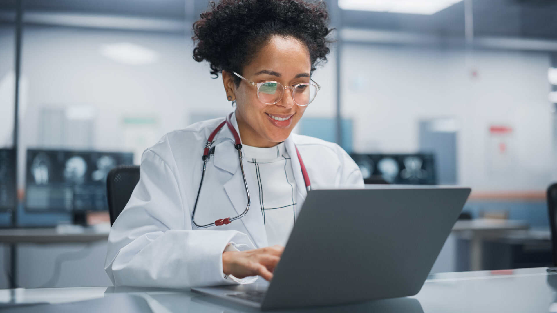Hospital Doctors Office: Portrait of Young Promising Black Physician Works on Laptop Computer. Female Medical Professional in White Lab Coat Looking for Patient Treatment While Sitting at Her Desk Hospital Doctors Office: Portrait of Young Promising Black Physician Works on Laptop Computer. Female Medical Professional in White Lab Coat Looking for Patient Treatment While Sitting at Her Desk
